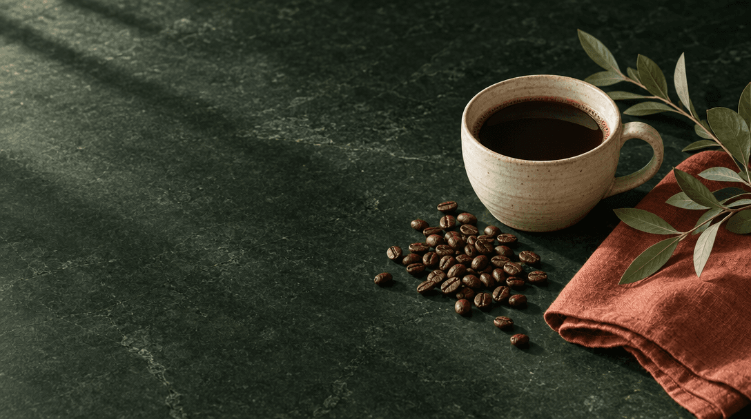 Cream ceramic coffee cup, roasted coffee beans and rust linen on a dark green stone surface